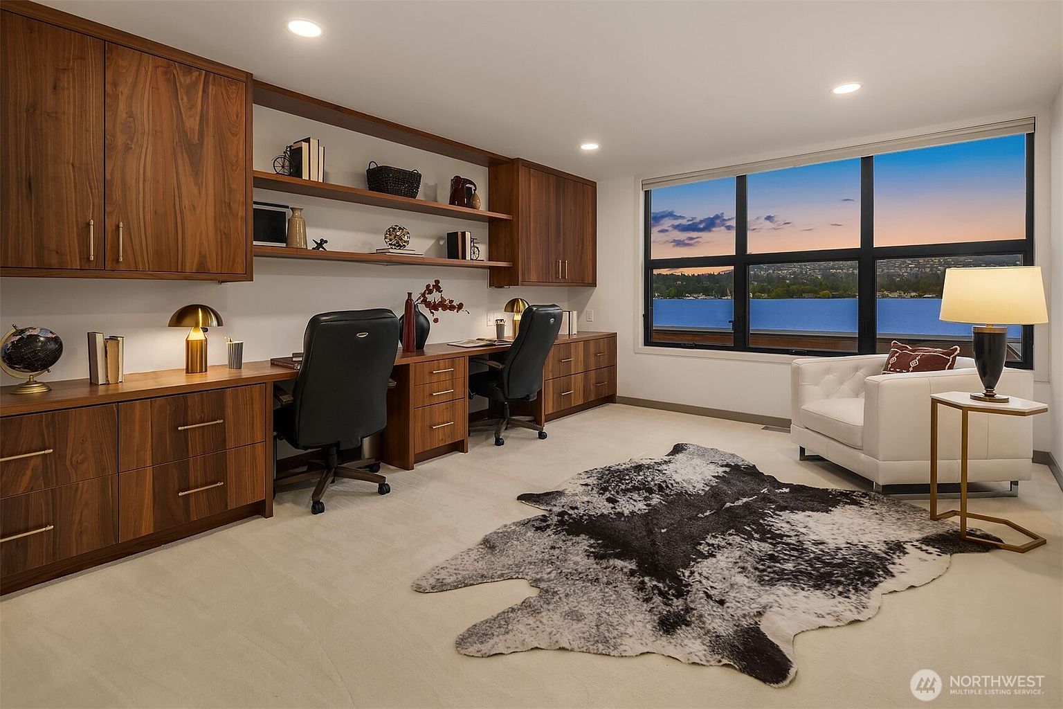 This sophisticated home office features custom built-in walnut cabinetry and shelving, providing ample storage and a sleek, professional aesthetic. The room is anchored by a dual-desk setup and a plush white armchair, all set against a neutral carpet and a striking cowhide rug. A large window offers a stunning, panoramic view of the water, creating a serene and inspiring workspace.