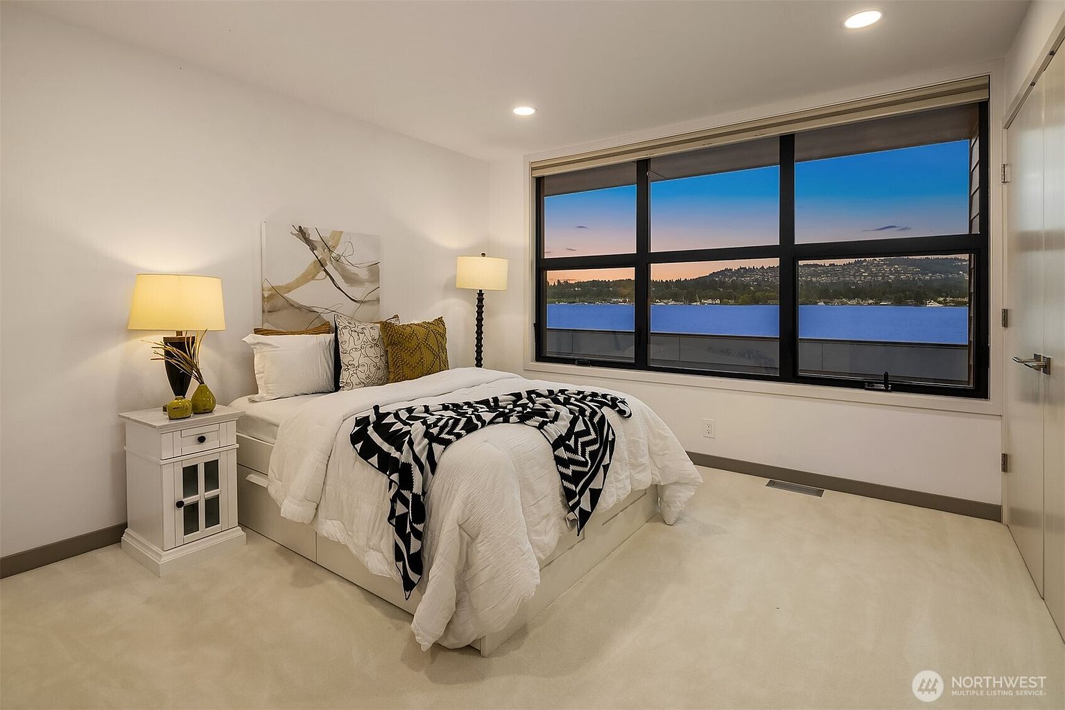This bright and modern bedroom features a comfortable bed dressed in white linens with a bold, patterned black-and-white throw blanket. A large, expansive window serves as the focal point, offering a stunning, unobstructed view of a serene lake at dusk. The room is minimalist in design, with neutral walls and carpet, creating a peaceful and inviting atmosphere.