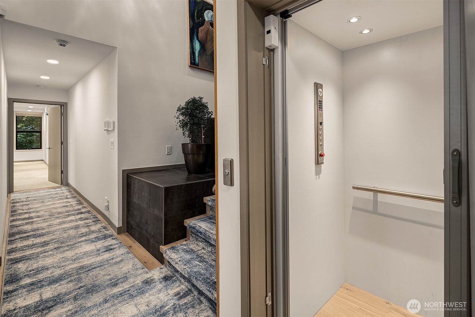 This interior view captures a modern hallway featuring a residential elevator on the right and a staircase leading upward on the left. The space is accented by a patterned runner rug, a dark tiled landing with a potted plant, and a piece of abstract art on the wall. The perspective highlights the clean, contemporary design and the seamless integration of accessibility features within the home's layout.