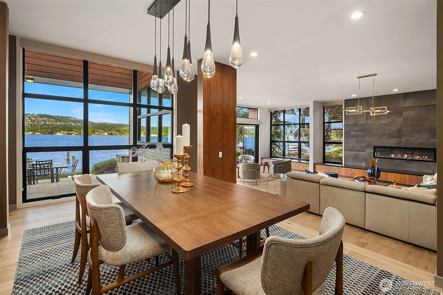 This elegant open-concept dining area features a rich wooden table surrounded by modern, upholstered chairs, set against a backdrop of floor-to-ceiling windows that offer a stunning panoramic view of the lake. A cluster of contemporary glass pendant lights hangs above the table, while the adjacent living room showcases a sleek fireplace and a large, comfortable sectional sofa. The space exudes a sophisticated, airy atmosphere with warm wood tones and a seamless connection to the natural landscape outside.
