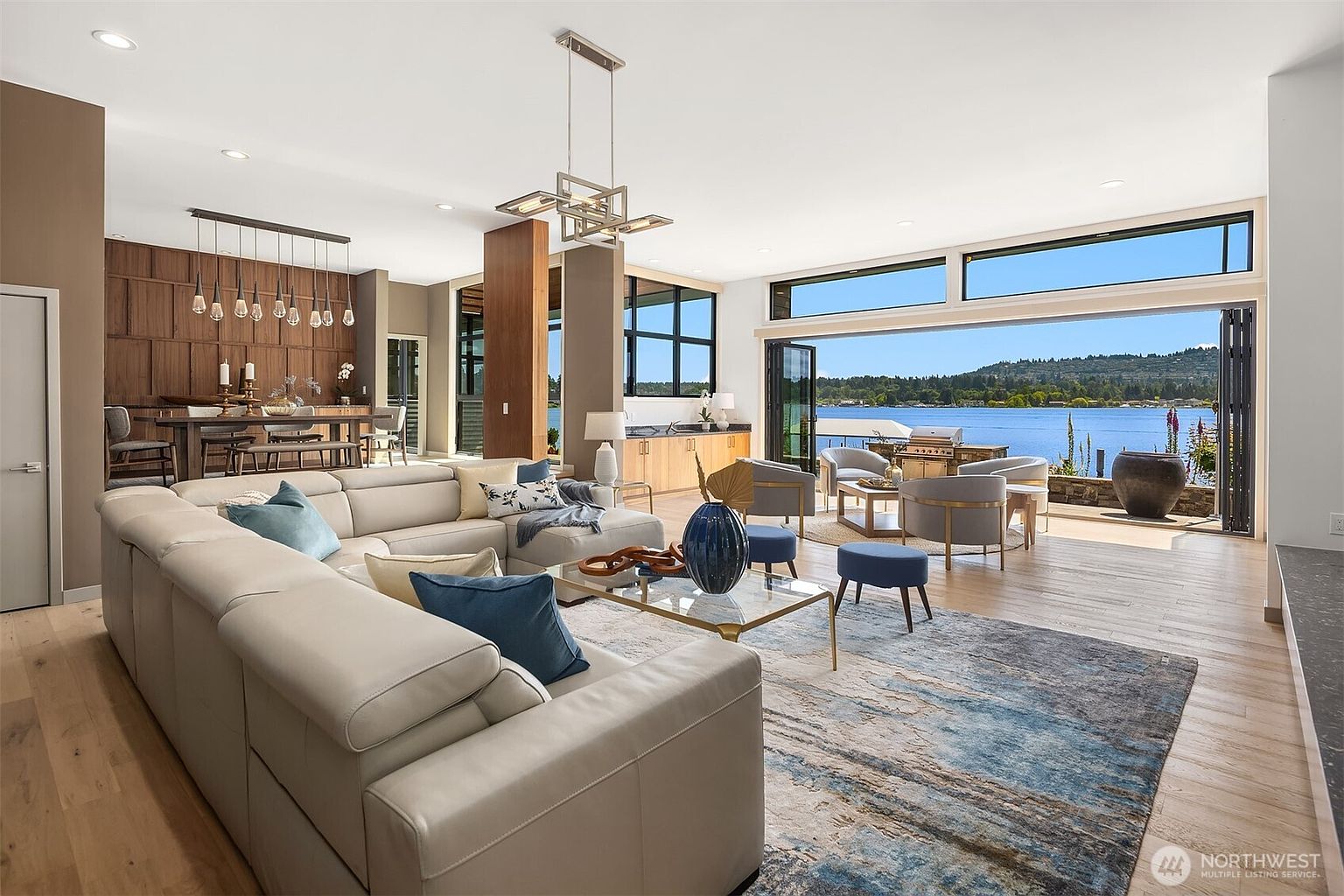 This expansive, modern living room features an open-concept layout that seamlessly transitions to a scenic outdoor patio overlooking a lake. The space is anchored by a large, light-colored sectional sofa, a contemporary glass-top coffee table, and a textured area rug, all illuminated by a striking geometric chandelier. With its floor-to-ceiling glass walls and sophisticated wood-paneled dining area in the background, the room exudes a luxurious, airy, and tranquil atmosphere.