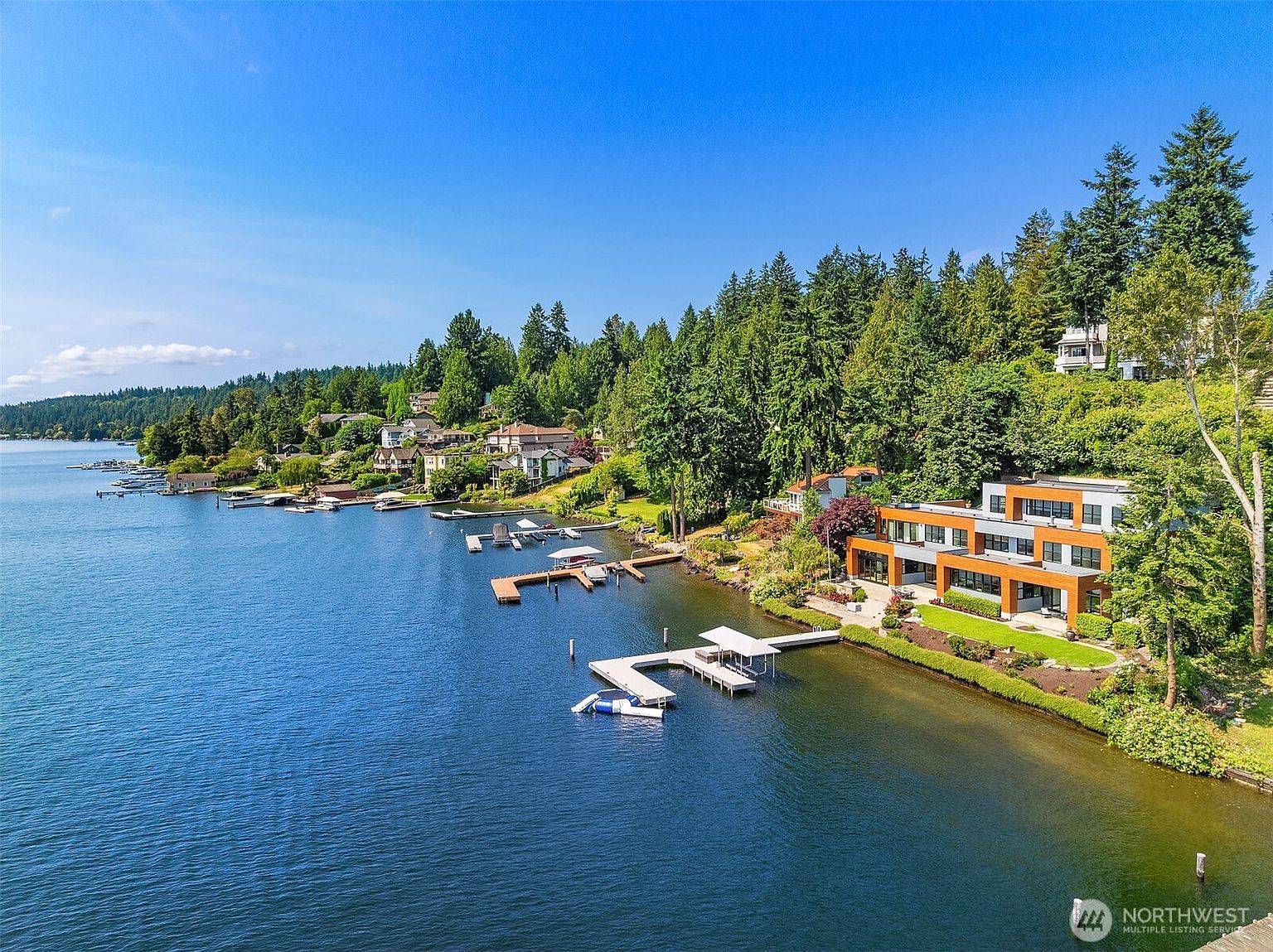 This high-angle aerial shot captures a stunning modern waterfront residence nestled along a lush, tree-lined shoreline. The multi-level home features contemporary architecture with warm wood accents and expansive glass windows, complemented by a private dock extending into the calm, deep blue water. The surrounding landscape is rich with mature evergreen trees, creating a serene and private atmosphere typical of a luxury lakeside retreat.