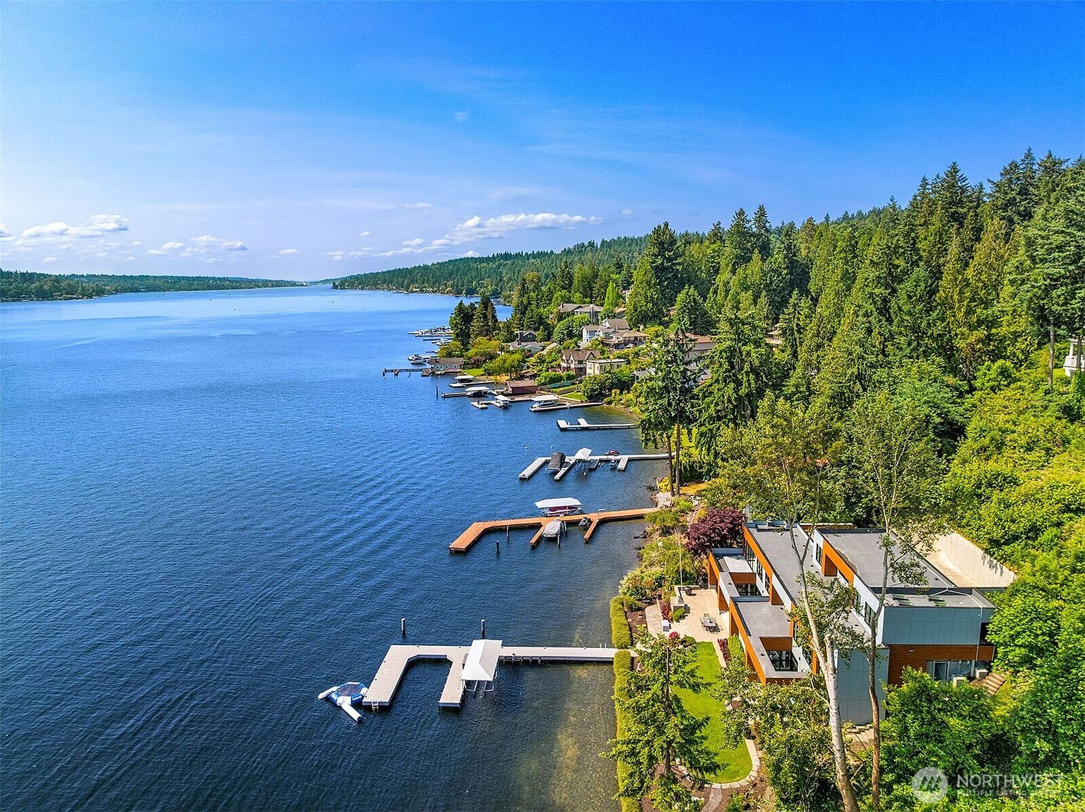 This stunning aerial view captures a modern, multi-level waterfront residence nestled along a lush, tree-lined shoreline. The property features a private dock extending into the expansive, deep blue lake, complemented by a contemporary architectural design with flat roofs and large windows. The surrounding landscape is heavily wooded, providing a serene and secluded atmosphere that highlights the home's prime location.