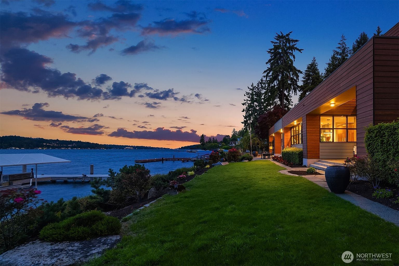 This stunning rear exterior view captures a modern, wood-clad home at twilight, perfectly positioned along a serene waterfront. The scene features a lush, manicured lawn leading to a private dock, all set against a dramatic, colorful sunset sky. The warm glow from the home's interior lighting creates an inviting and luxurious atmosphere, highlighting the seamless transition between indoor living and the natural landscape.
