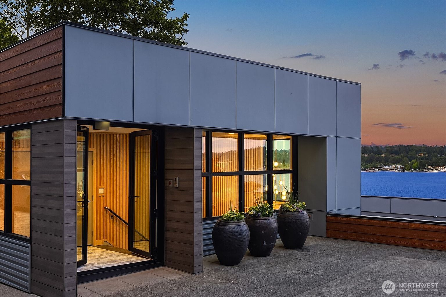 This modern rooftop terrace features a sleek, contemporary design with a combination of wood-paneled and smooth grey siding. The space includes a glass-paned entryway leading into a warm, wood-accented interior, complemented by three large, dark decorative planters. The perspective captures a stunning sunset view over a calm body of water, emphasizing the luxurious and serene outdoor living experience.