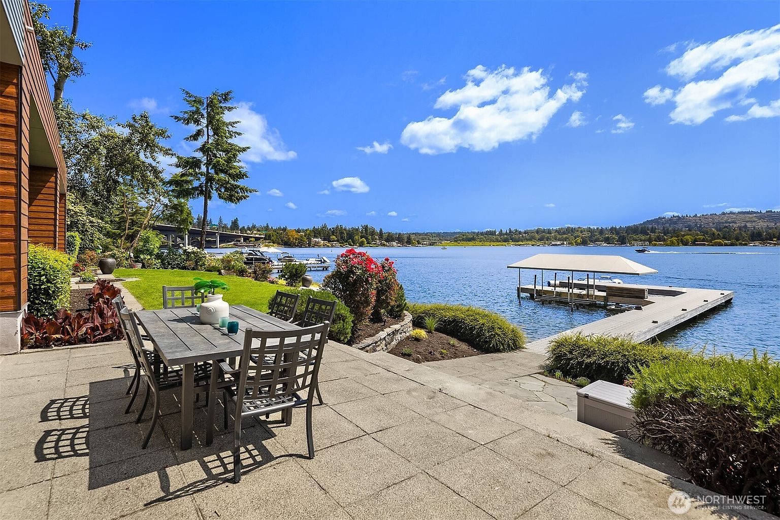 This serene waterfront patio features a modern outdoor dining set positioned on large stone pavers, overlooking a calm lake with a private dock and boat lift. The scene is framed by lush landscaping, including vibrant red flowers and manicured shrubs, with a clear blue sky overhead. The perspective captures a peaceful, high-end outdoor living space that seamlessly blends architectural design with natural beauty.