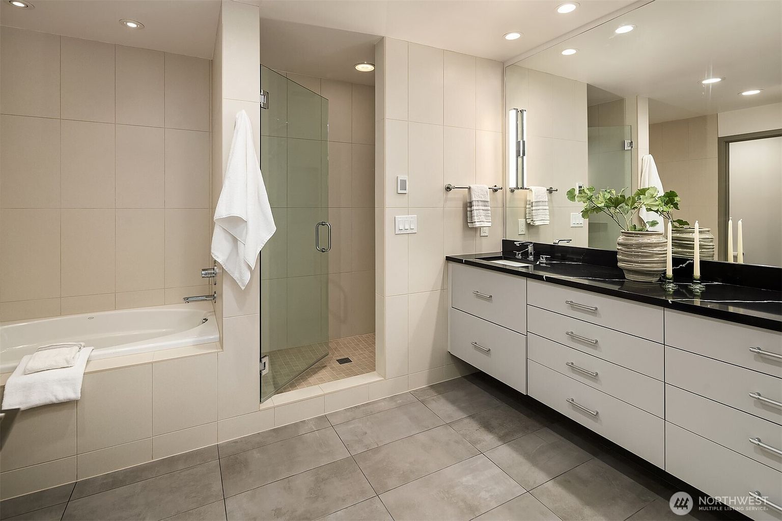 This modern primary bathroom features a sleek, minimalist design with large-format light-colored wall tiles and dark grey floor tiles. The space includes a deep soaking tub, a glass-enclosed walk-in shower, and a long vanity with a black countertop and double sinks. The perspective is a wide-angle shot that captures the clean lines and contemporary aesthetic of the entire room.