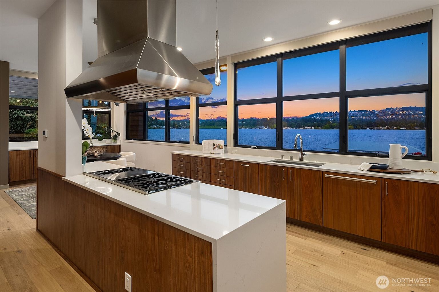 This modern, high-end kitchen features warm wood cabinetry paired with sleek white quartz countertops and a stainless steel range hood. A large window wall provides a stunning, panoramic view of the water at sunset, creating a serene and luxurious atmosphere. The open-concept layout seamlessly integrates the cooking area with the living space, emphasizing a bright and airy aesthetic.