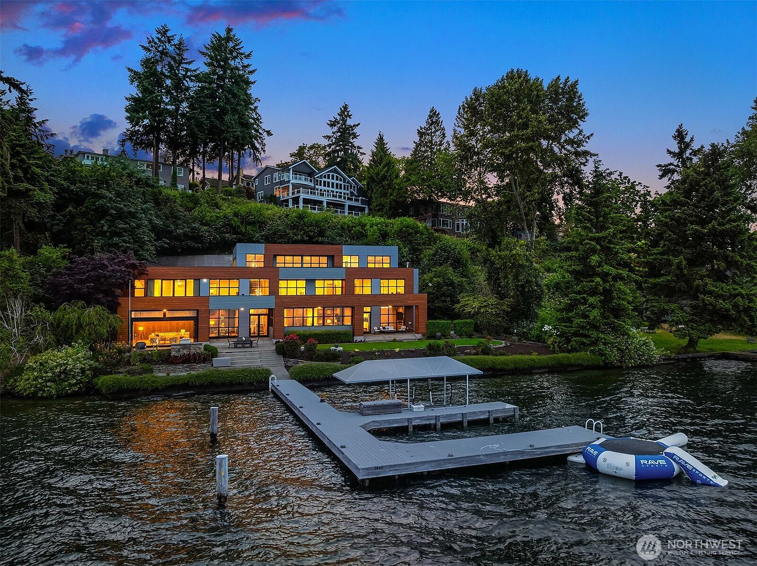 This stunning contemporary waterfront estate is captured from a low-angle perspective across the water, showcasing its multi-level architectural design and expansive glass walls. The property features a private dock with a boat lift, a manicured lawn, and a seamless transition between the indoor living spaces and the outdoor terrace. The twilight sky provides a dramatic backdrop, highlighting the warm interior lighting and the sophisticated, modern aesthetic of the home.