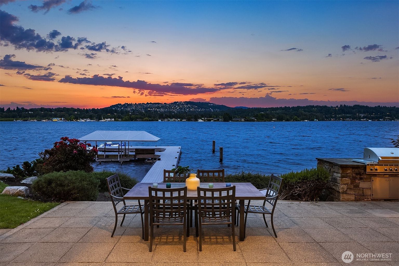 This stunning waterfront patio offers a serene outdoor dining space featuring a metal table and chairs set against a breathtaking sunset over the lake. The scene includes a private dock with a boat lift, lush landscaping, and a built-in outdoor grill, creating an ideal setting for evening relaxation or entertaining. The perspective captures the seamless transition from the paved patio to the expansive water view, emphasizing the property's prime location.
