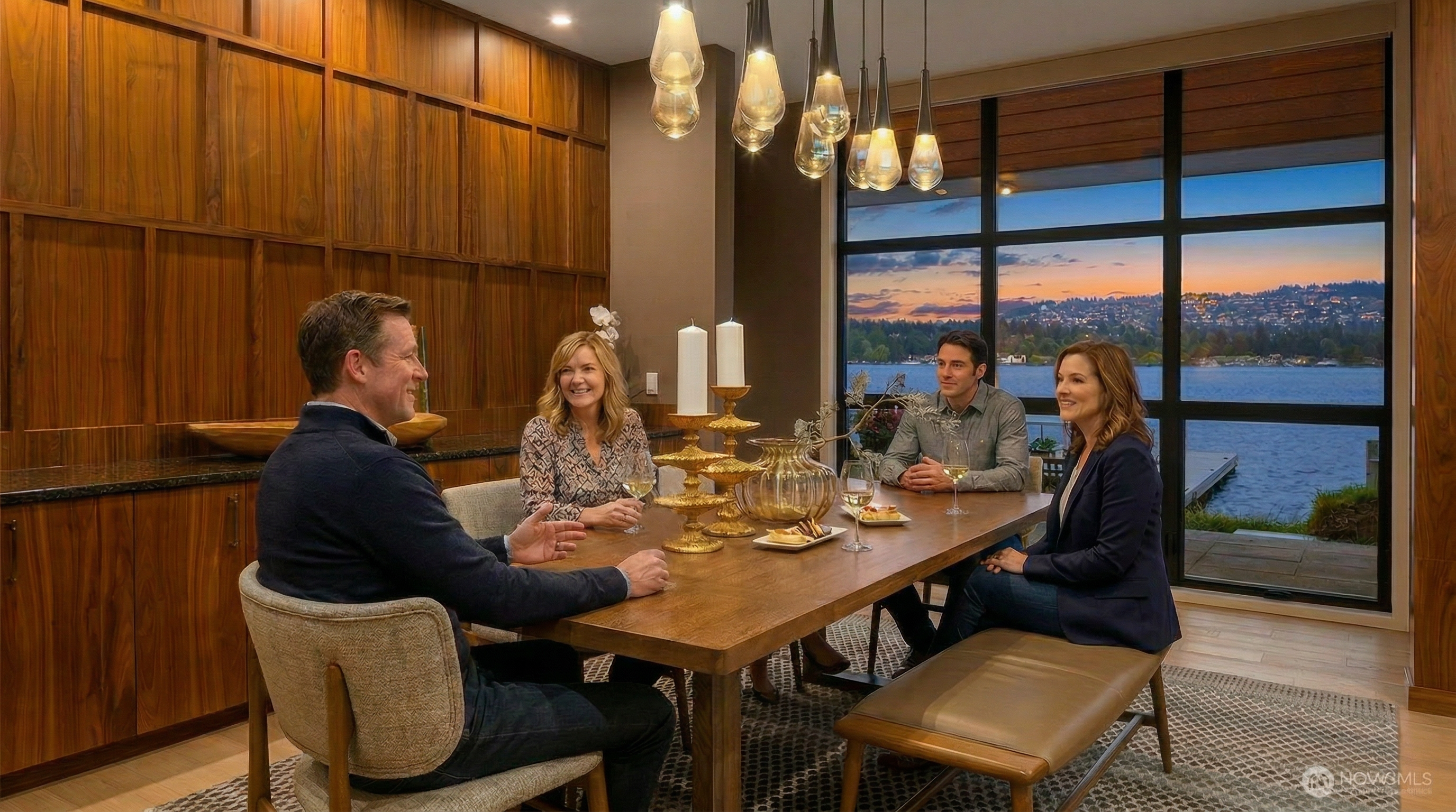 This elegant dining room features a warm, wood-paneled accent wall and a large wooden table paired with upholstered chairs and a leather bench. A striking cluster of teardrop-shaped pendant lights hangs above the table, while floor-to-ceiling windows provide a breathtaking, panoramic view of a lake at sunset. The space exudes a sophisticated, modern aesthetic, perfectly blending natural textures with a serene waterfront backdrop.