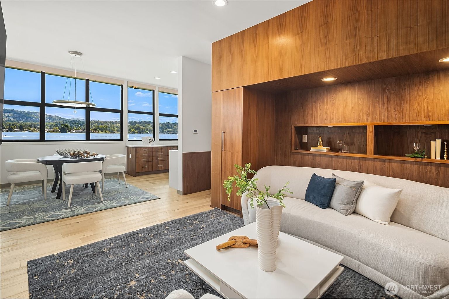 This sophisticated living space features a modern, open-concept design with a warm wood-paneled accent wall and a plush, light-colored sofa. The room seamlessly transitions into a dining area with floor-to-ceiling windows that offer a stunning, expansive view of the lake and surrounding landscape. The combination of natural wood textures, contemporary furniture, and abundant natural light creates an inviting and luxurious atmosphere.