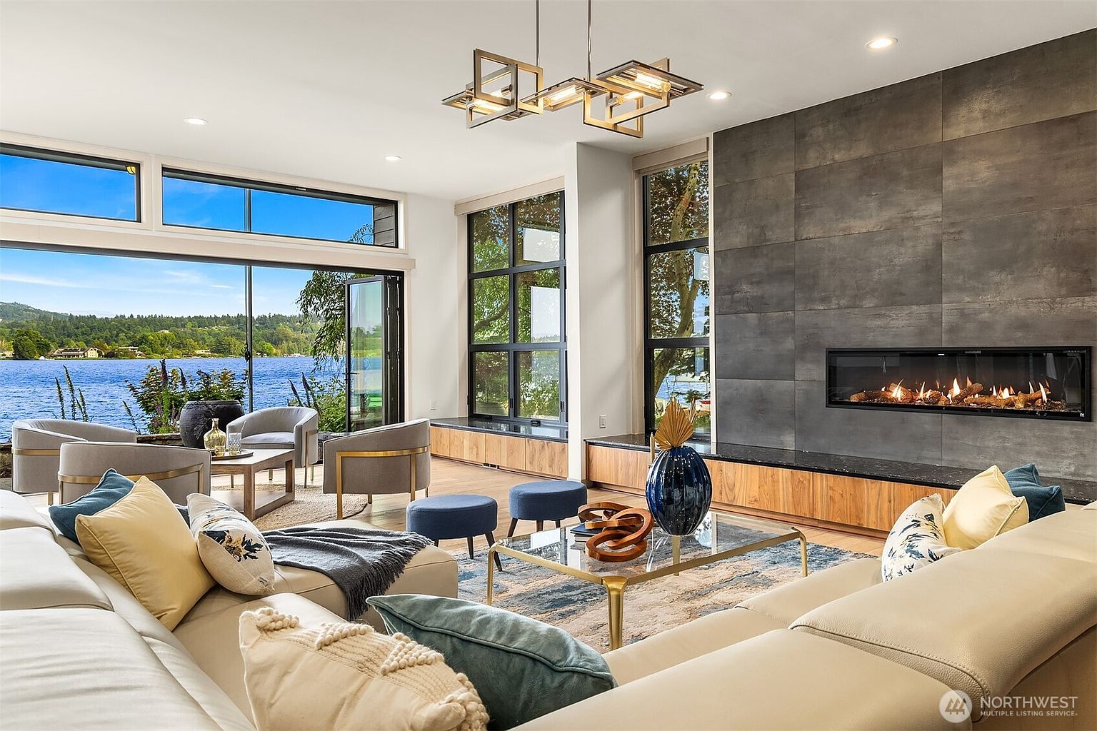 This sophisticated living room features a contemporary design with a large, light-colored sectional sofa and a sleek, linear fireplace set into a dark, textured accent wall. Expansive floor-to-ceiling windows provide a stunning, unobstructed view of the lake, seamlessly blending indoor comfort with the natural beauty of the outdoors. The space is accented by a modern geometric chandelier, a glass-topped coffee table, and rich wood cabinetry that runs along the base of the windows.