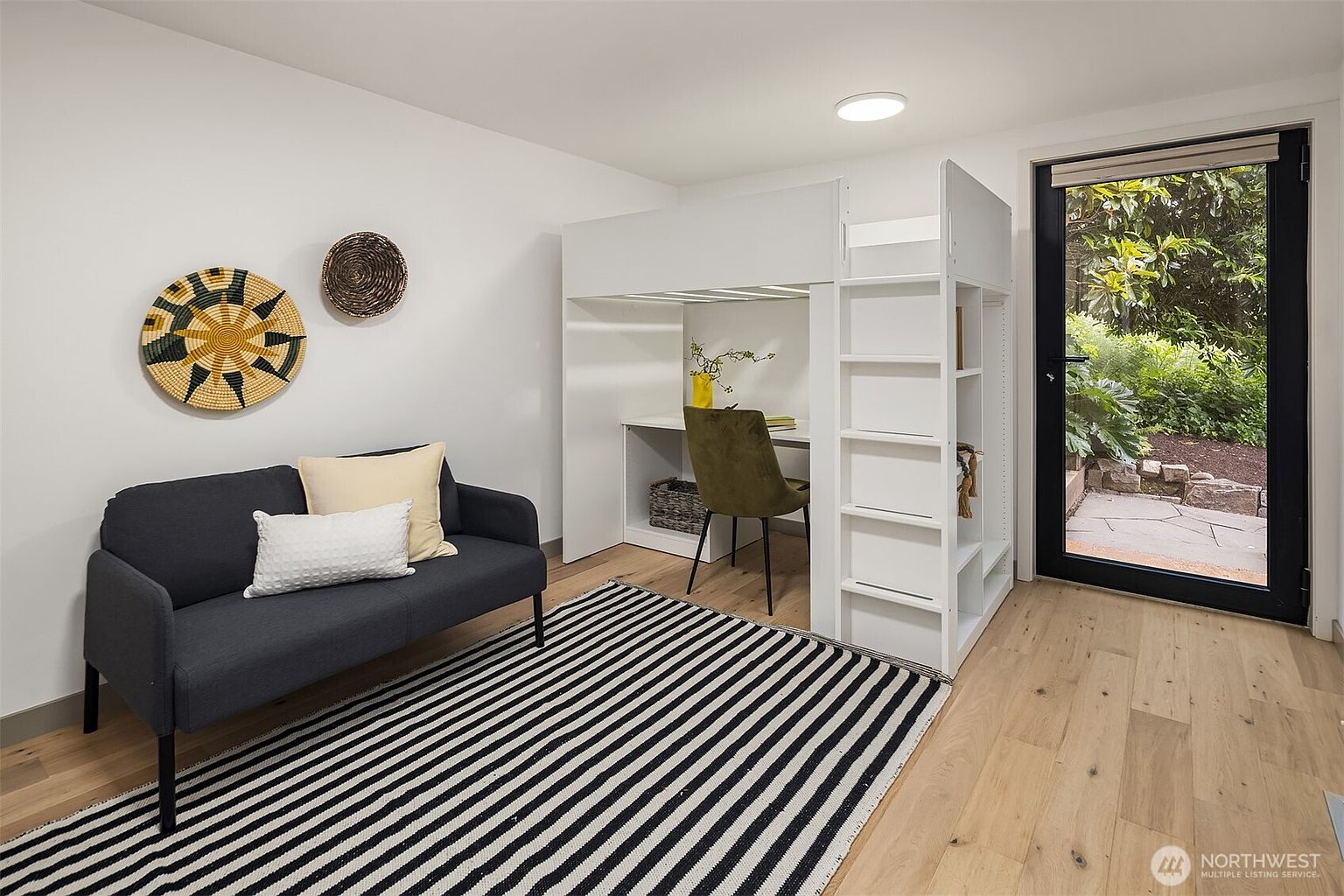 This versatile room features a modern white loft bed with an integrated desk and shelving unit, maximizing space efficiency. A dark gray sofa sits on a bold black-and-white striped rug, creating a cozy seating area, while a glass door provides natural light and a view of the lush greenery outside. The room's clean lines and neutral color palette offer a contemporary and functional aesthetic suitable for a bedroom or home office.