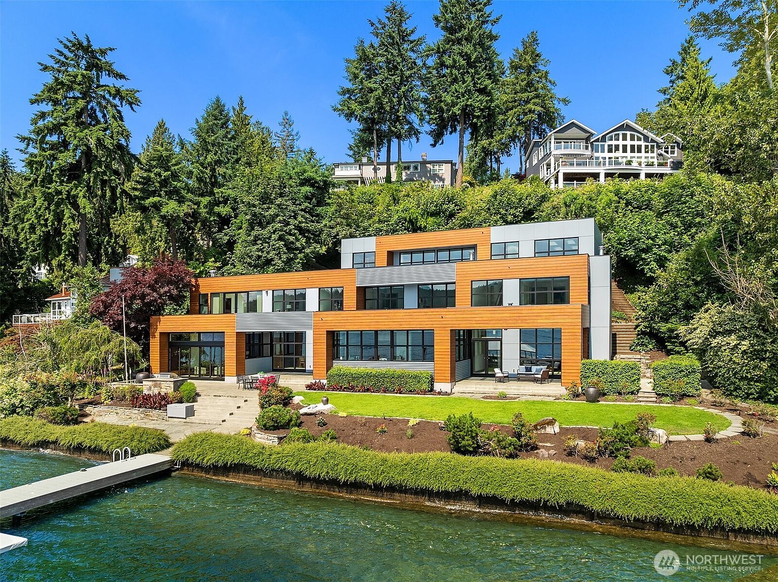 This stunning modern waterfront estate features a multi-level architectural design with a blend of warm wood siding and sleek gray panels. The home is nestled against a lush, tree-covered hillside and overlooks a serene lake, complete with a private dock and a manicured lawn. The expansive windows and tiered terraces emphasize a seamless connection between the luxurious interior and the natural landscape.