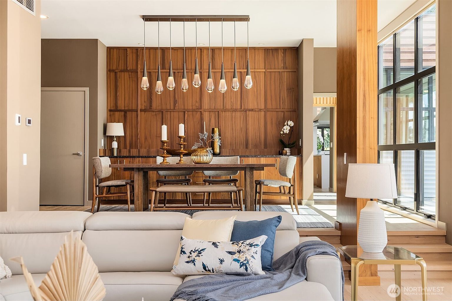 This elegant dining area features a warm, wood-paneled accent wall that serves as a sophisticated backdrop for a rustic wooden table and modern upholstered chairs. A row of contemporary pendant lights hangs above the table, casting a soft glow over the space, while a plush, light-colored sofa in the foreground anchors the open-concept living area. The room exudes a refined, transitional style, blending natural wood textures with clean lines and comfortable, inviting furnishings.