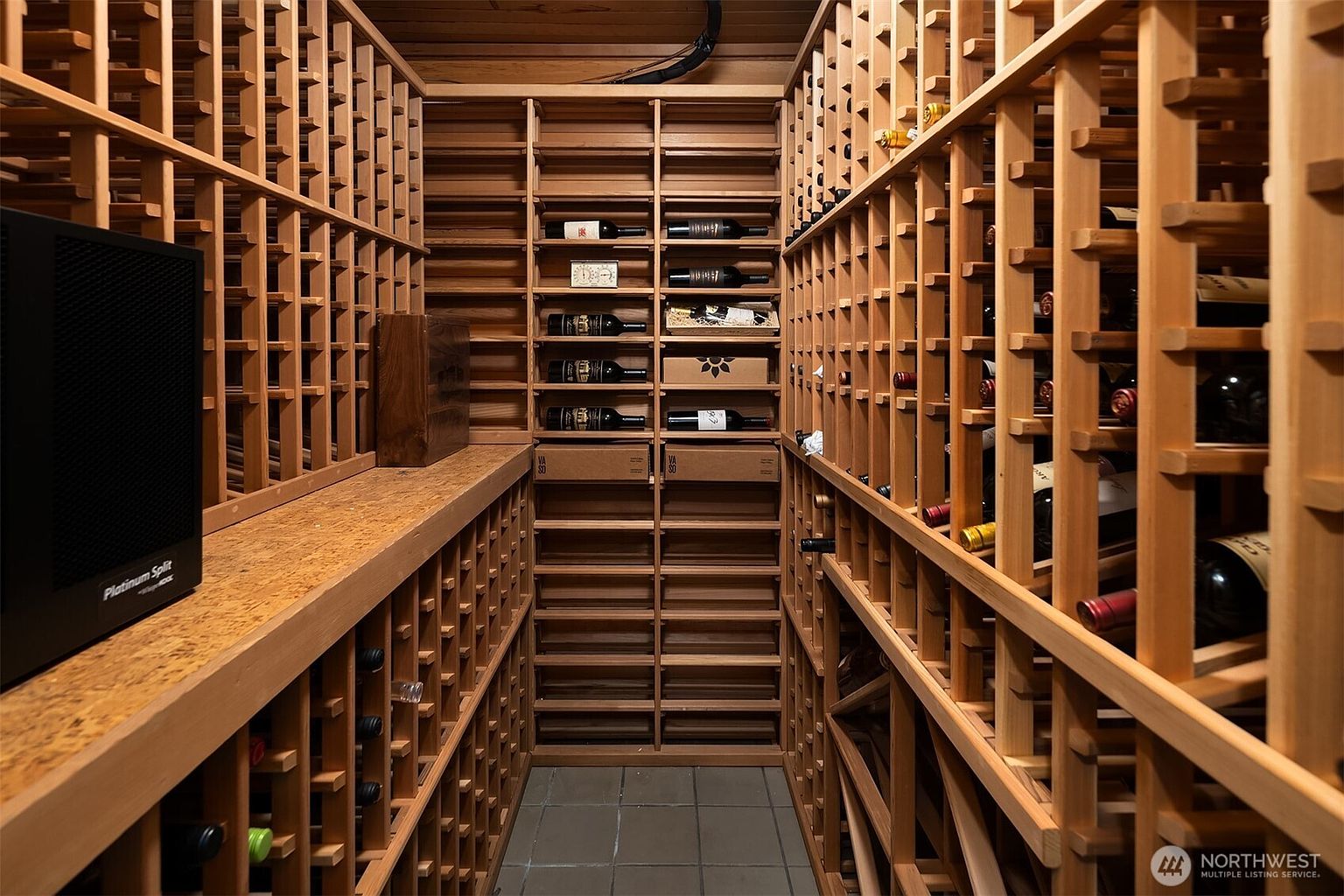 This dedicated wine cellar features extensive custom wooden racking systems designed for optimal bottle storage, creating a sophisticated and organized atmosphere. The space is climate-controlled with a visible Platinum Split cooling unit, emphasizing its functionality for serious wine collectors. The perspective is a centered, eye-level shot looking down the narrow aisle, highlighting the depth and craftsmanship of the wood shelving.