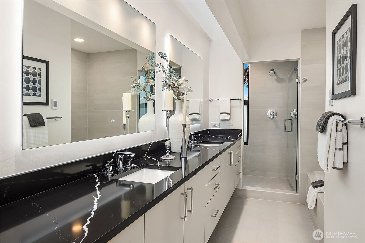 This modern primary bathroom features a sleek double vanity with a striking black marble countertop and white cabinetry. A large, illuminated mirror spans the wall, reflecting the clean lines of the space, while a glass-enclosed walk-in shower with light-toned tile adds to the contemporary aesthetic. The room is accented with elegant decor, including a tall vase and framed wall art, creating a sophisticated and spa-like atmosphere.