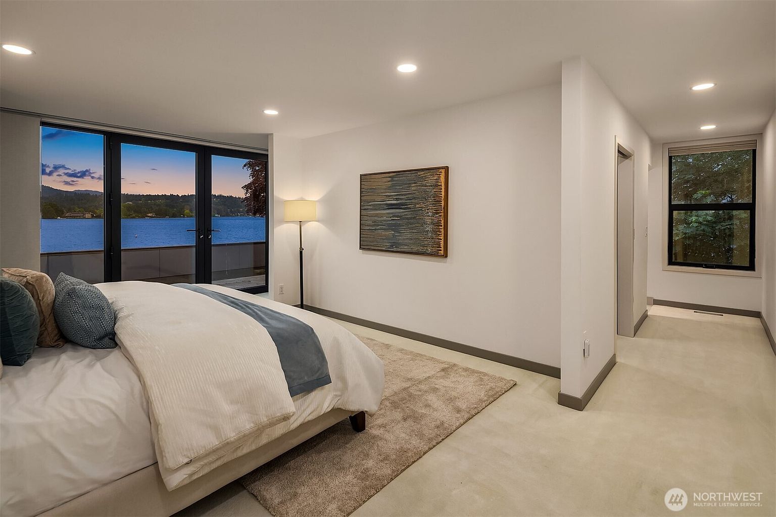 This primary bedroom features a serene, modern aesthetic with a plush bed dressed in neutral tones and a soft area rug. A large glass door provides a stunning, unobstructed view of the lake at sunset, while recessed lighting and a floor lamp create a warm, inviting atmosphere. The clean lines and minimalist decor emphasize the tranquil waterfront setting.