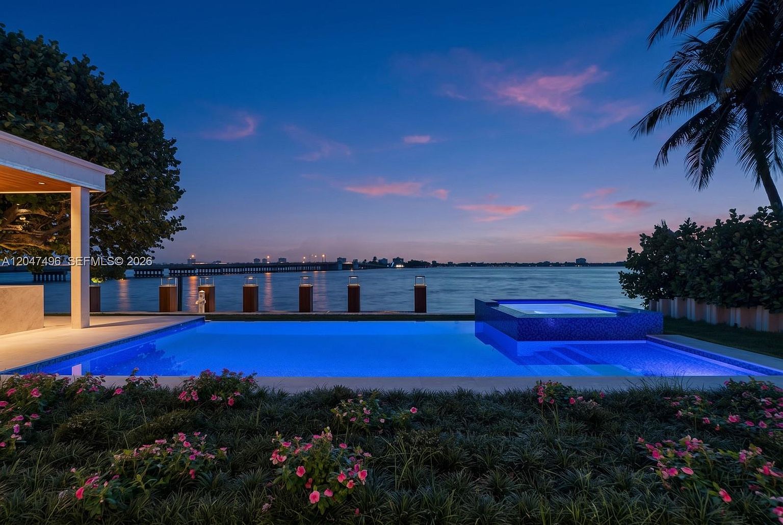 This stunning waterfront property features a modern infinity-edge pool and integrated spa, illuminated with vibrant blue lighting against a twilight sky. The pool deck overlooks a calm bay with a long pier extending into the water, framed by a lush foreground of flowering plants and a silhouetted palm tree. The cinematic perspective captures the serene transition from the manicured garden to the expansive horizon, emphasizing the luxurious outdoor living space.