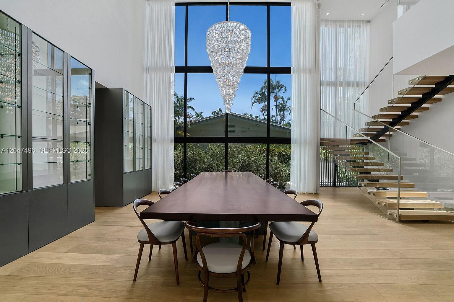 This elegant dining room features a long, dark wood table surrounded by modern chairs, set beneath a dramatic, multi-tiered crystal chandelier. The space is defined by expansive floor-to-ceiling windows that provide a clear view of the outdoors, complemented by tall, sheer white curtains. To the left, a sleek, dark-framed glass display cabinet adds a sophisticated touch, while a floating staircase with glass railings ascends on the right, emphasizing the room's airy, double-height ceiling.