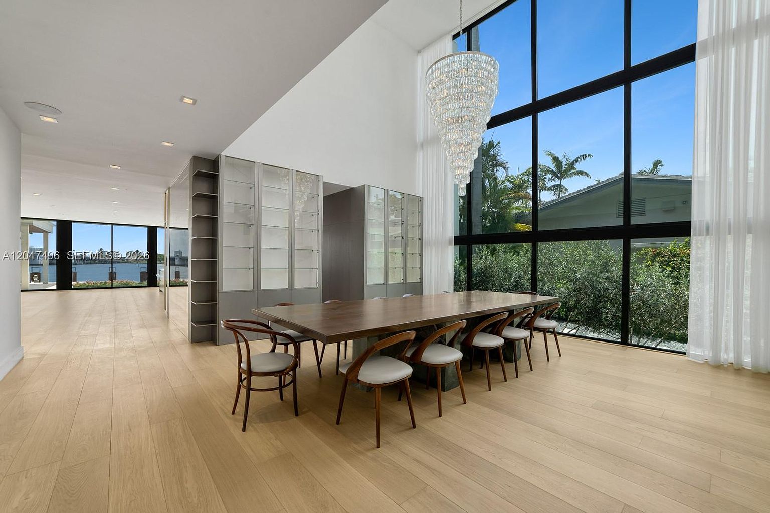This spacious dining room features a long, dark wood table surrounded by ten modern chairs with curved backs and light-colored cushions. A grand, multi-tiered crystal chandelier hangs above the table, serving as a focal point against the backdrop of expansive, floor-to-ceiling windows that offer views of the lush greenery outside. The room is finished with light wood flooring and includes a sleek, built-in storage unit with glass-fronted cabinets, creating an elegant and airy atmosphere.