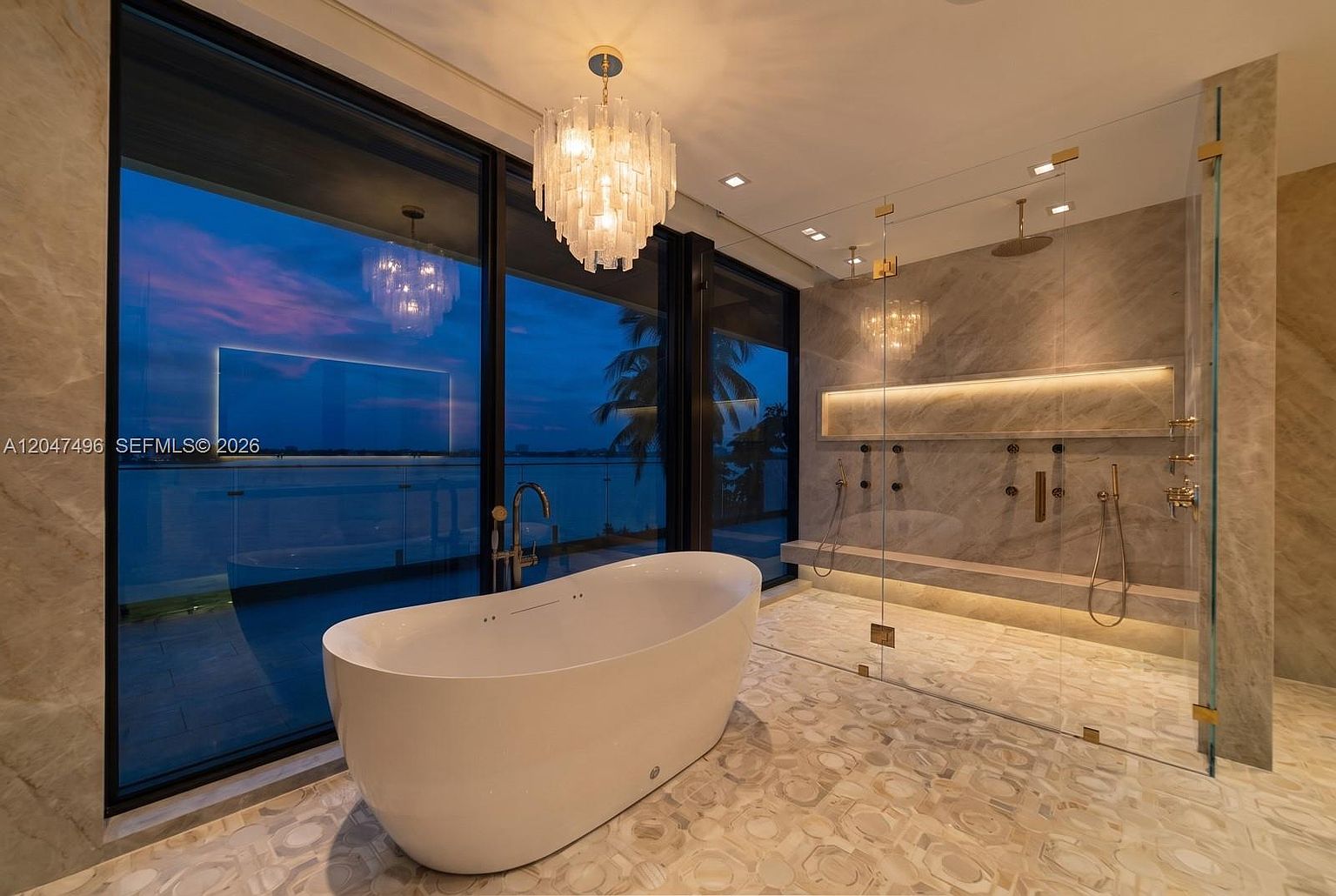 This luxurious primary bathroom features a sleek, freestanding soaking tub positioned in front of expansive floor-to-ceiling windows that offer a stunning twilight view of the water. To the right, a spacious walk-in shower is enclosed by glass and finished with elegant, light-toned stone tile and integrated ambient lighting. The room is illuminated by a sophisticated crystal chandelier, creating an atmosphere of refined elegance and modern tranquility.