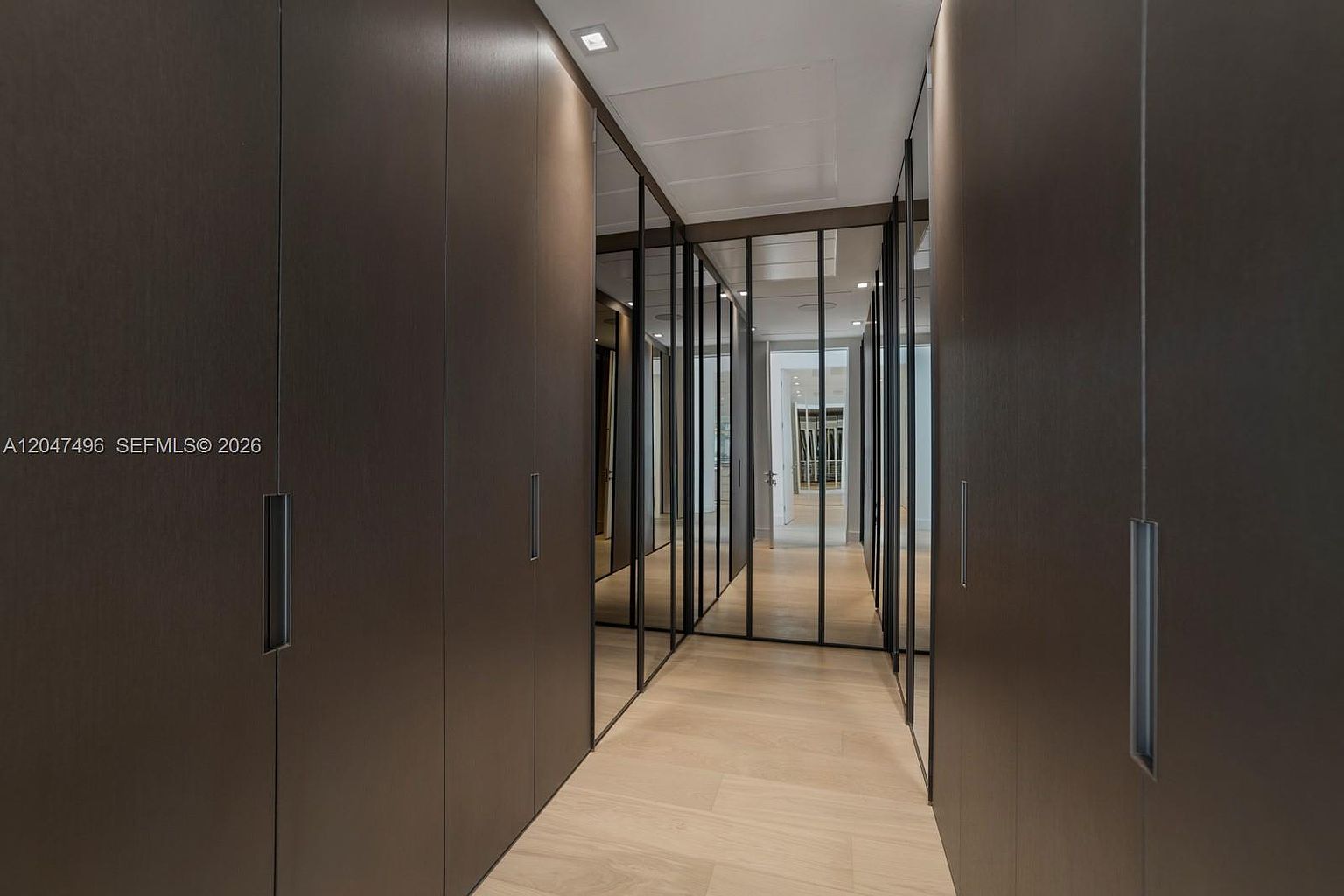This sleek, modern walk-in closet features dark, matte-finish cabinetry lining both sides of a narrow corridor. The space is accented by tall, mirrored panels with thin black frames that add depth and a contemporary aesthetic. Light-toned wood flooring runs throughout, leading the eye toward a bright doorway at the end of the hall.