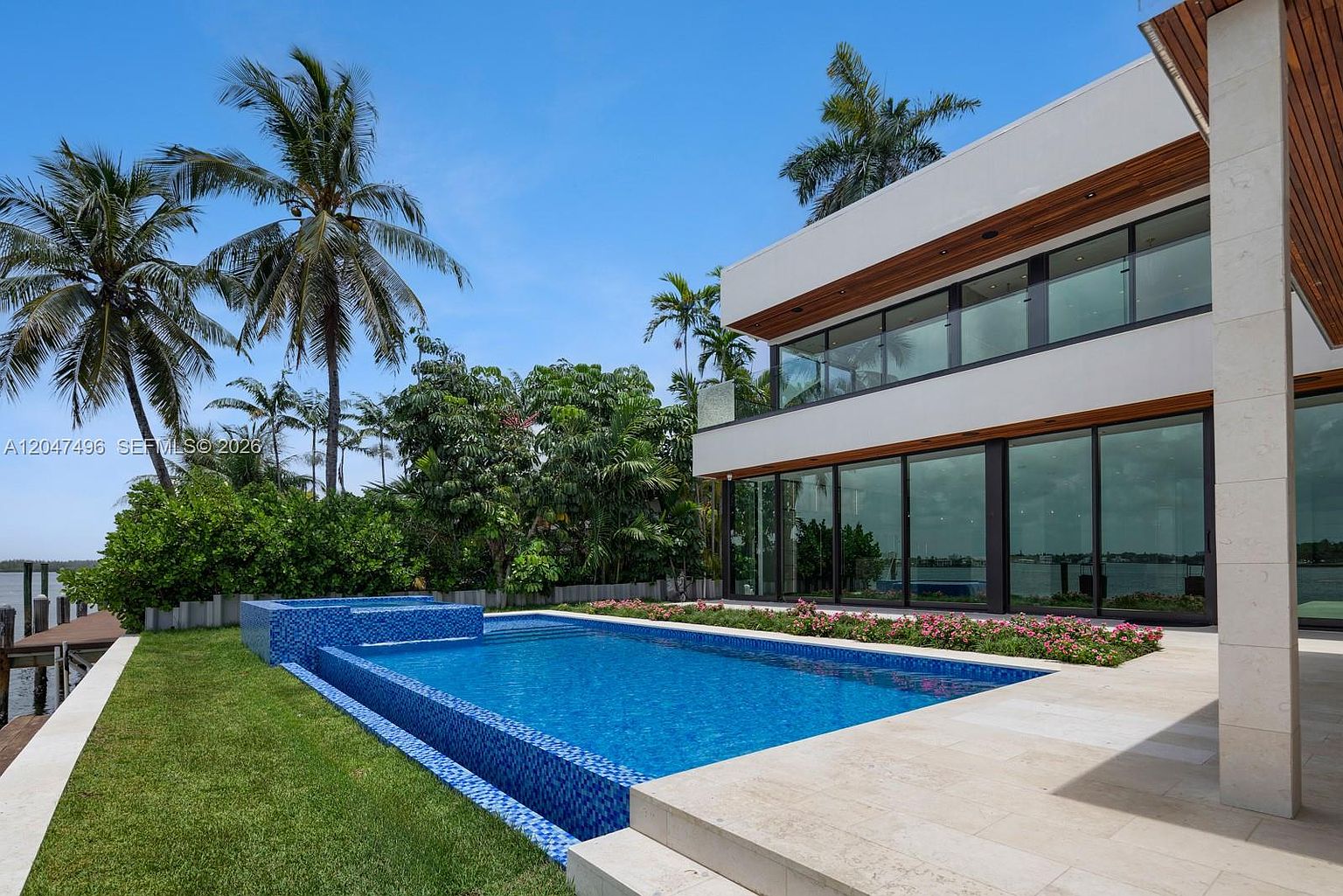 This modern, two-story waterfront residence features a sleek architectural design with expansive glass walls that offer seamless indoor-outdoor living. A vibrant blue tiled pool and integrated spa sit prominently in the foreground, set against a backdrop of lush tropical palm trees and a manicured lawn. The clean lines of the structure and the serene water view create a sophisticated, resort-like atmosphere.