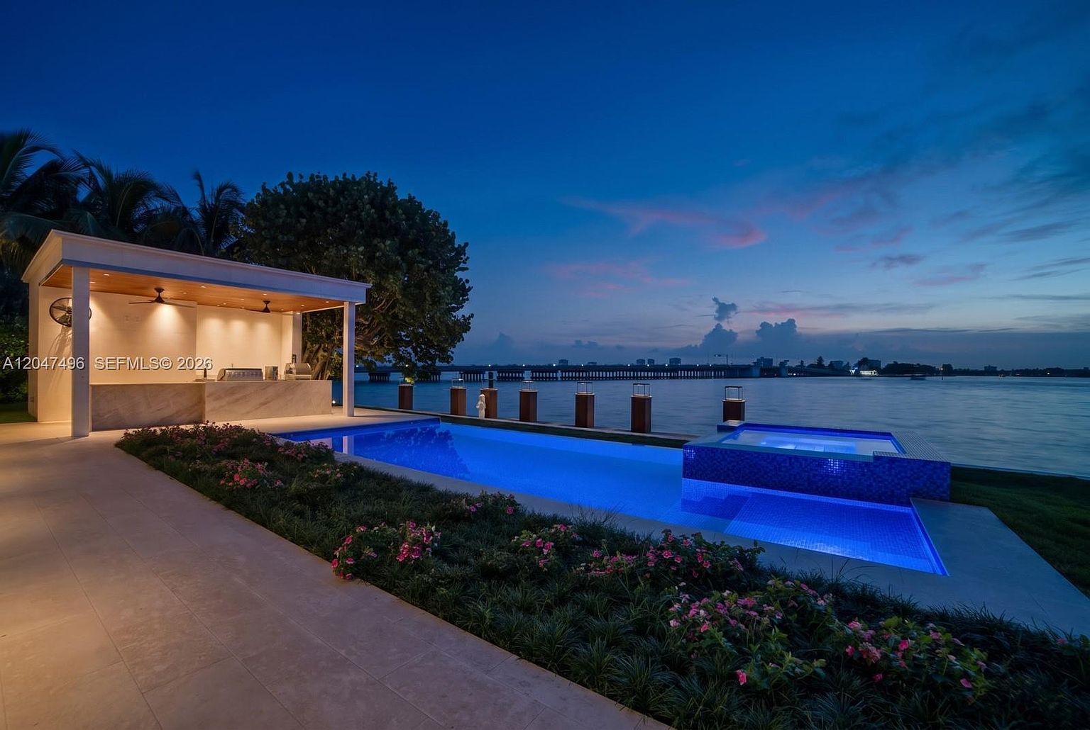 This stunning waterfront property features a sleek, rectangular infinity-edge pool and an elevated spa, both illuminated with vibrant blue lighting at dusk. Adjacent to the pool is a covered outdoor kitchen area with a stone-topped bar and ceiling fans, perfect for entertaining. The scene is set against a tranquil bay view with a distant bridge, creating a sophisticated and serene atmosphere.