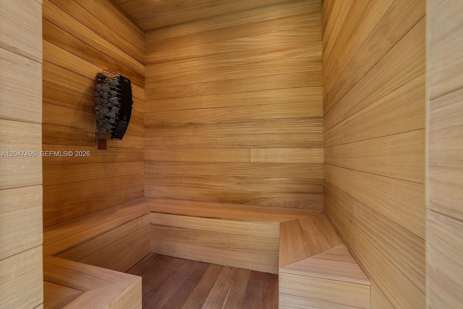 This image shows a compact, custom-built sauna constructed entirely from light-toned wood paneling. The space features built-in wooden benches arranged in an L-shape and a wall-mounted electric sauna heater filled with dark stones. The perspective is from the doorway, looking into the warm, minimalist interior.