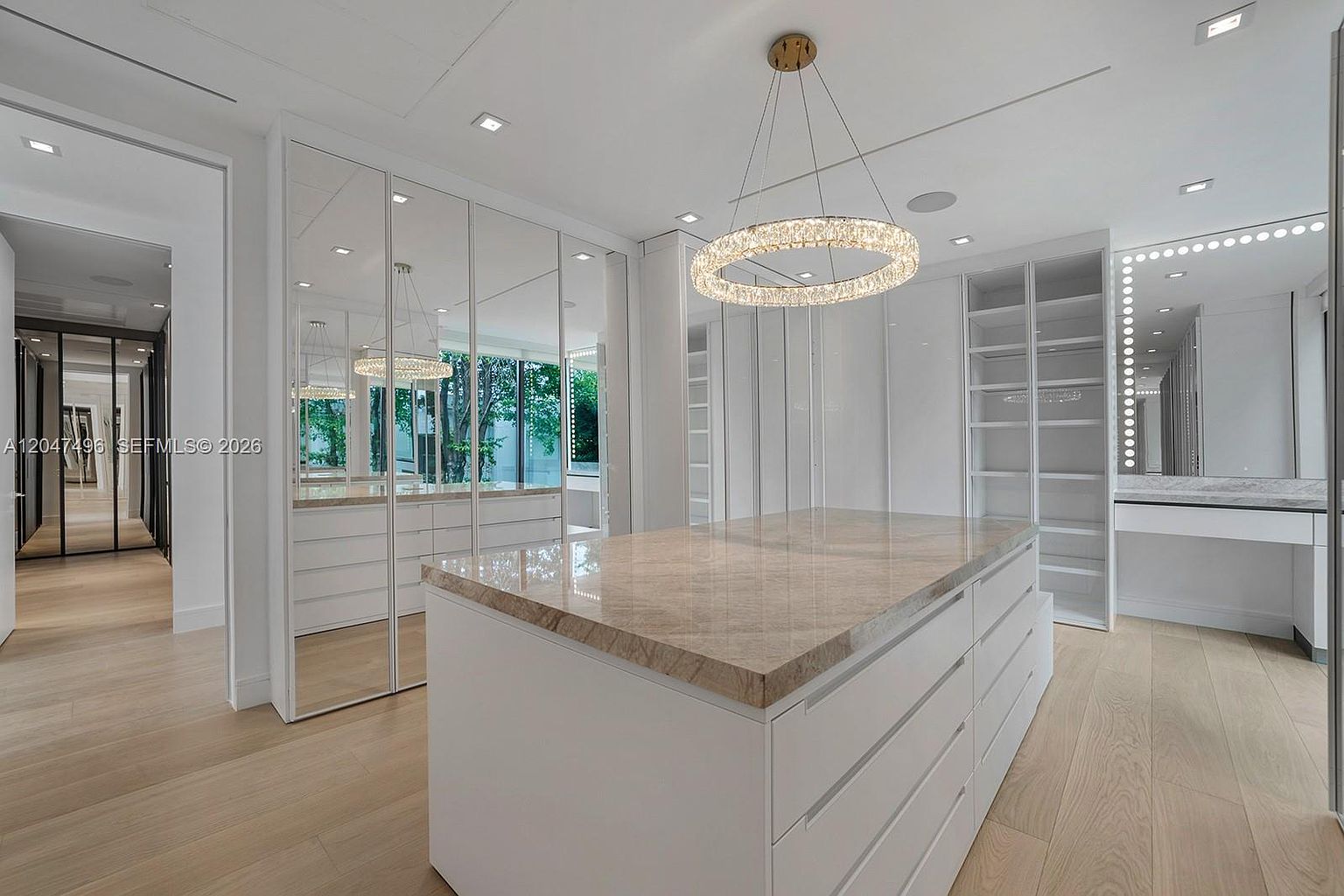 This luxurious walk-in closet features a central island with a stone countertop and ample drawer storage, surrounded by white cabinetry and mirrored wardrobe doors. A modern circular crystal chandelier serves as a focal point, while a dedicated vanity area with a lighted mirror adds functionality. The space is finished with light wood flooring, creating a bright, sophisticated, and organized atmosphere.