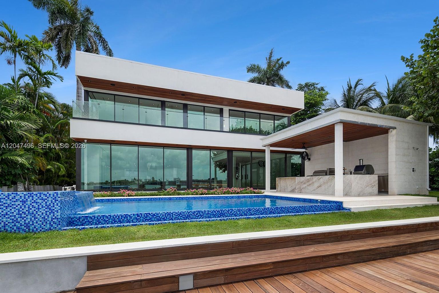 This modern, two-story residence features a striking contemporary design with extensive glass walls and clean, horizontal lines. The backyard is highlighted by a rectangular swimming pool with a built-in water feature, adjacent to a covered outdoor kitchen area with a stone-topped island. The foreground includes a wooden deck, creating a seamless transition between the indoor living spaces and the lush, tropical landscaping.