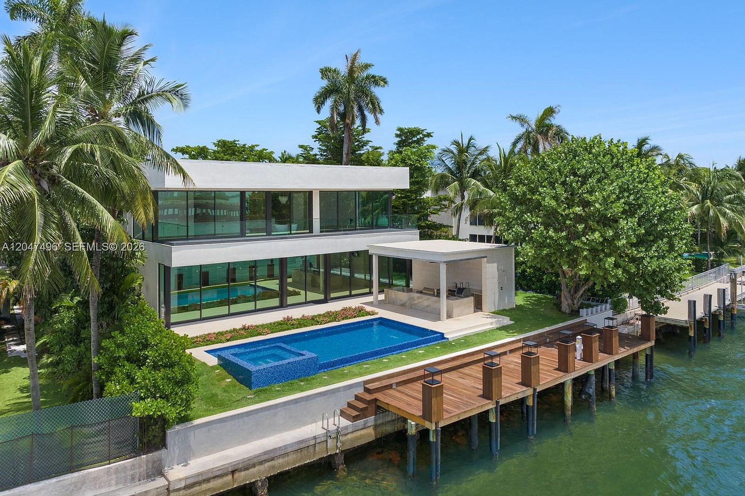 This modern waterfront estate features a sleek, two-story design with extensive glass walls that provide seamless indoor-outdoor living. The backyard includes a rectangular swimming pool with an integrated spa, a covered outdoor kitchen area, and a wooden dock extending into the water. Lush tropical landscaping and tall palm trees frame the property, creating a luxurious and serene coastal atmosphere.