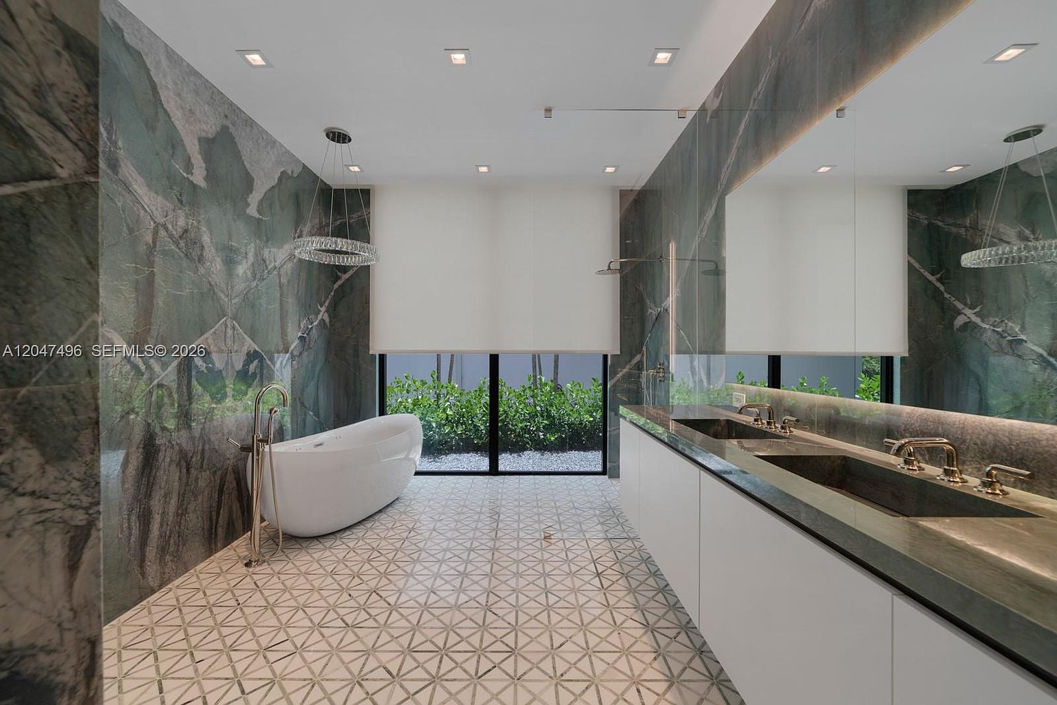 This luxurious primary bathroom features a striking, dark-veined stone wall that serves as a dramatic backdrop for a sleek, freestanding soaking tub. A long, dual-sink vanity with a stone countertop runs along the right wall, complemented by a large mirror and modern fixtures. The space is finished with geometric patterned floor tiles and large windows that provide a view of the lush greenery outside.