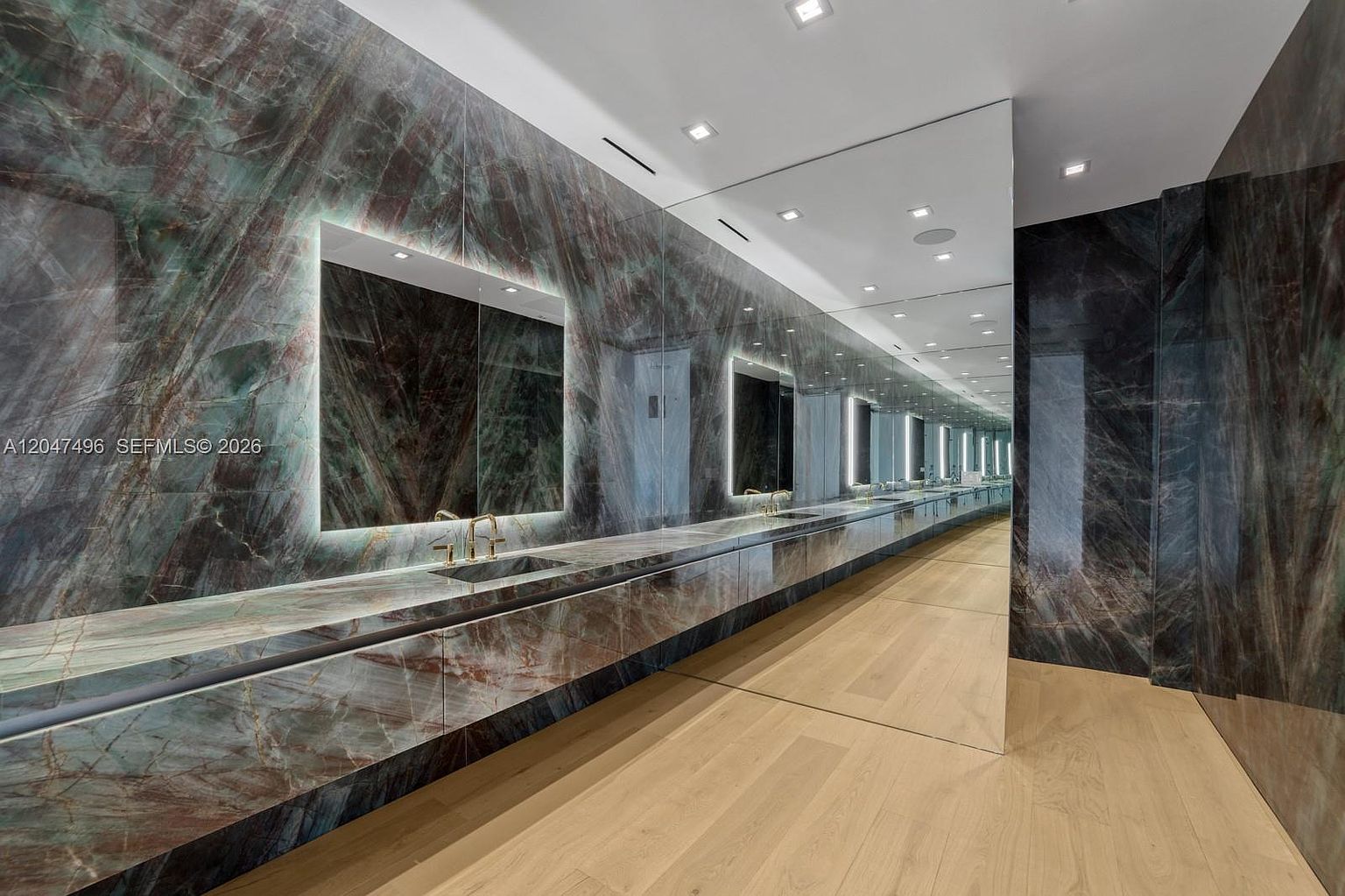 This expansive, ultra-modern bathroom features a long, floating vanity clad in dramatic, dark-veined stone that extends across the wall. The space is defined by a massive, continuous mirror that reflects the room, creating an illusion of infinite depth, complemented by integrated LED lighting behind the vanity mirrors. Light-toned wood flooring provides a warm contrast to the dark stone, while recessed ceiling lights illuminate the sleek, minimalist design.