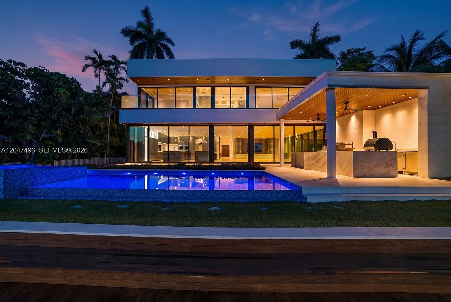 This modern two-story residence is captured at dusk, showcasing a sleek architectural design with expansive glass walls that illuminate the interior. The rear exterior features a large swimming pool with vibrant blue lighting, adjacent to a covered outdoor kitchen area equipped with a grill and stone-topped island. The scene is framed by lush tropical landscaping and palm trees, creating a sophisticated and inviting atmosphere for evening entertaining.
