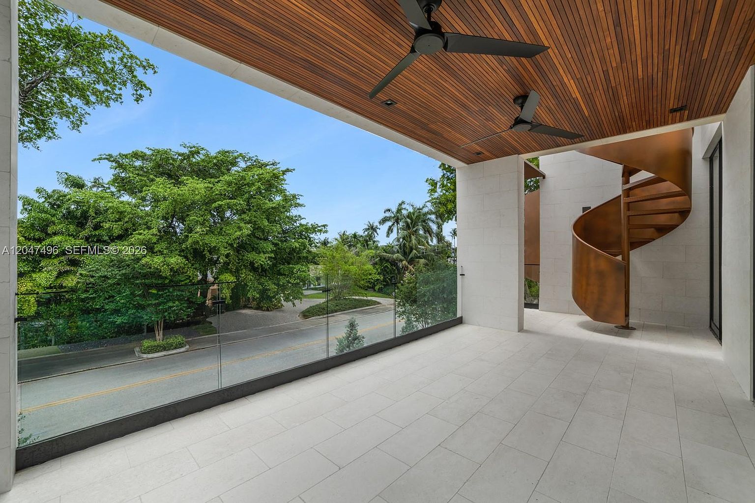 This modern, covered balcony features a sleek glass railing that provides an unobstructed view of the street and lush surrounding trees. The space is defined by a warm wood-slatted ceiling with two integrated ceiling fans and a striking, sculptural spiral staircase finished in a metallic bronze tone. The light-colored stone flooring creates a clean, minimalist aesthetic that emphasizes the open-air design.