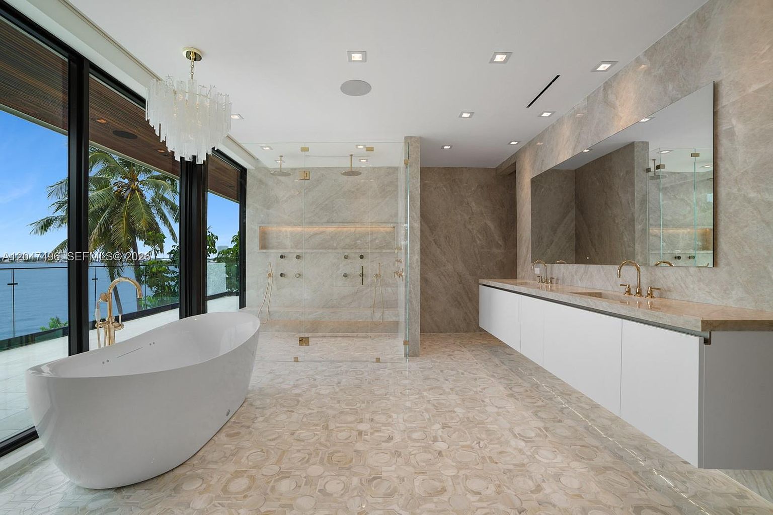 This luxurious primary bathroom features a sleek, freestanding soaking tub positioned next to expansive floor-to-ceiling windows that offer a panoramic ocean view. The space is finished with light-toned stone flooring and wall cladding, complemented by a long, minimalist white vanity with dual gold-toned faucets and a large, frameless mirror. A spacious, glass-enclosed walk-in shower with dual rainfall showerheads and a recessed niche completes the high-end, contemporary design.