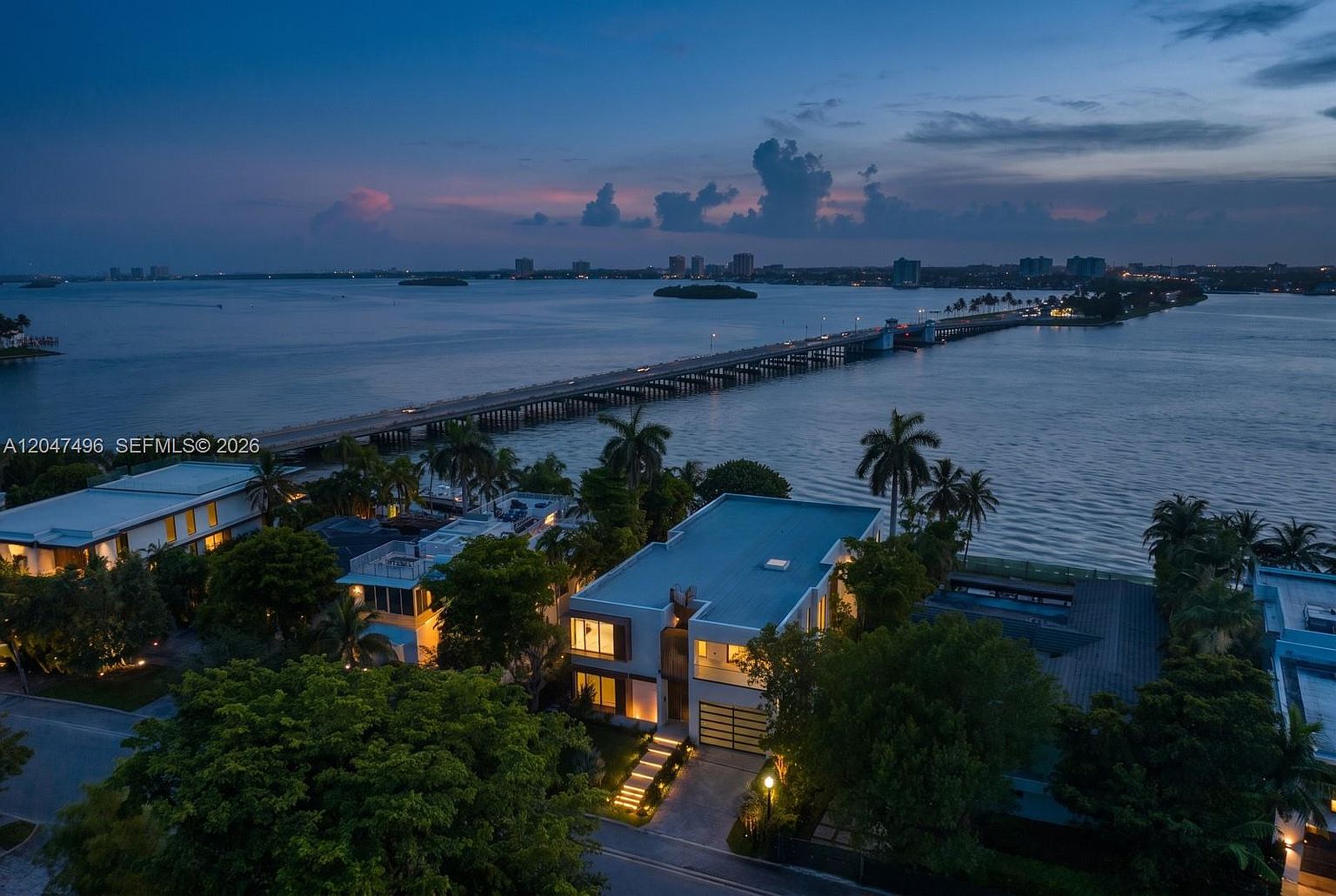 This high-angle aerial shot captures a contemporary waterfront residence at twilight, showcasing its modern architectural design and proximity to a bridge spanning the bay. The property features a multi-level structure with illuminated windows, a paved driveway with integrated landscape lighting, and lush surrounding greenery. The expansive water view and the distant city skyline create a serene and luxurious atmosphere.
