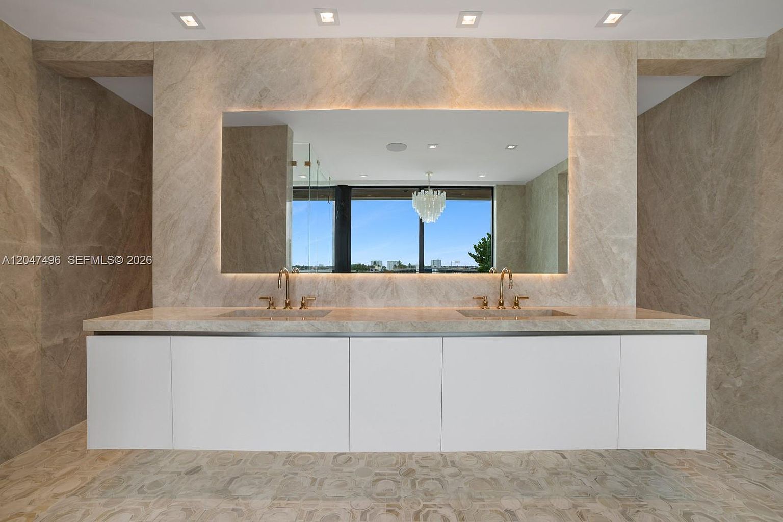 This luxurious primary bathroom features a double vanity with a long, light-toned stone countertop and integrated sinks, set against a wall clad in large-format, veined stone tiles. A wide, backlit mirror spans the width of the vanity, reflecting a chandelier and a large window that provides a view of the outdoors. The floor is covered in a distinctive, circular-patterned stone tile, creating a sophisticated and cohesive aesthetic.