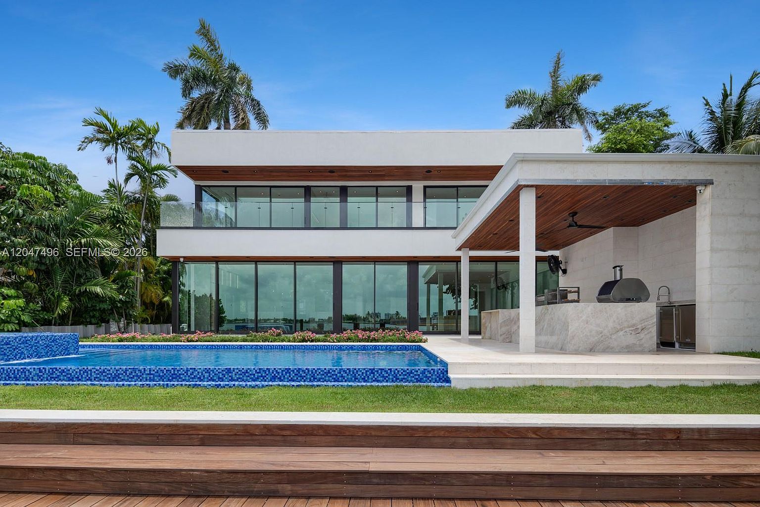 This modern, two-story residence features a sleek design with extensive floor-to-ceiling glass walls that seamlessly connect the indoor living spaces to the outdoor area. The backyard includes a rectangular swimming pool with blue mosaic tiles and a covered patio area equipped with an outdoor kitchen and a pizza oven. The property is framed by lush palm trees and a manicured lawn, creating a sophisticated and tranquil waterfront atmosphere.