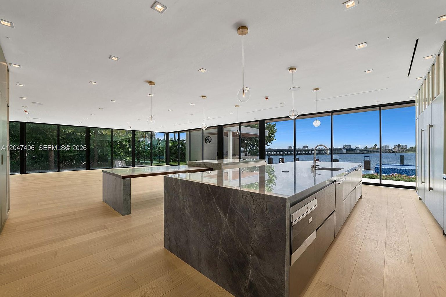 This expansive, modern kitchen features a large central island with a stone countertop and integrated appliances, complemented by a secondary attached dining or prep surface. The room is defined by floor-to-ceiling glass walls that provide panoramic views of the waterfront and pool area outside. Light wood flooring runs throughout the space, which is illuminated by a series of minimalist pendant lights and recessed ceiling fixtures.