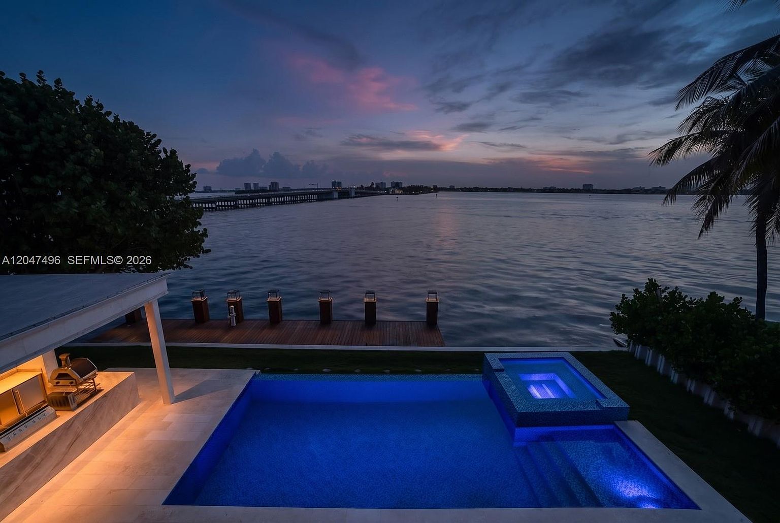 This elevated perspective captures a luxurious waterfront pool and spa area at twilight, featuring vibrant blue underwater lighting. A wooden dock extends into the calm bay, which is framed by a distant bridge and city skyline under a soft, colorful sunset sky. To the left, a covered outdoor kitchen area with a built-in grill provides a sophisticated space for entertaining.