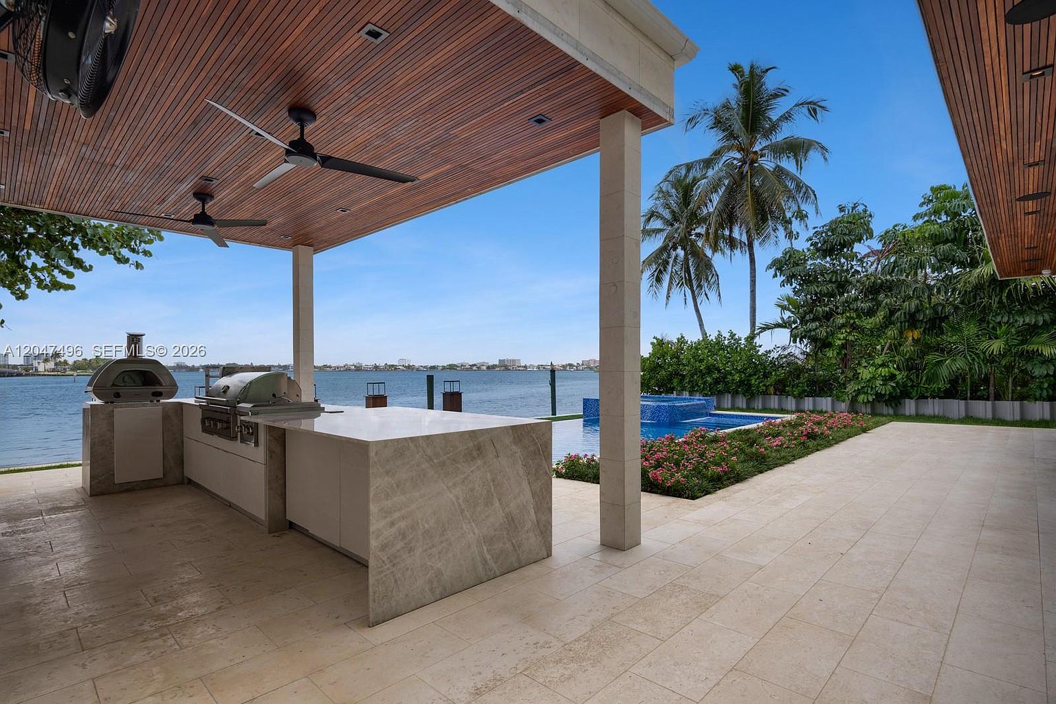 This expansive covered outdoor living space features a sleek, stone-topped kitchen island equipped with a built-in grill and a pizza oven. The area is sheltered by a warm-toned wood-paneled ceiling with integrated fans, overlooking a private swimming pool and a serene waterfront view. The scene is framed by lush tropical landscaping and a clear blue sky, creating a luxurious and inviting atmosphere for outdoor entertaining.