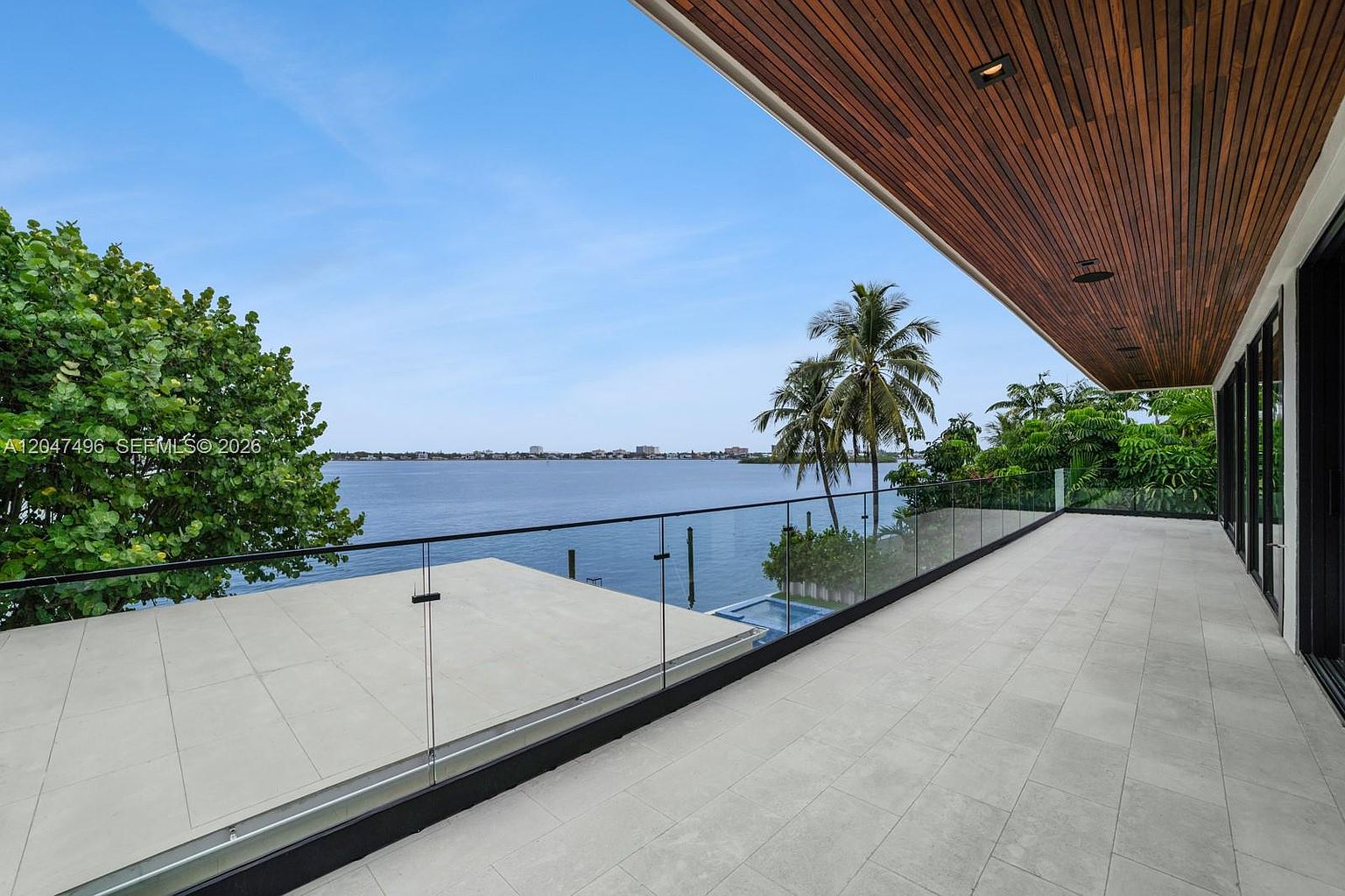 This expansive, modern balcony features light-toned tile flooring and a sleek glass railing that provides an unobstructed view of the water. The ceiling is finished with warm-toned wood slats, adding a sophisticated architectural element to the outdoor space. The perspective captures the serene waterfront setting, highlighting the seamless transition between the indoor living area and the scenic exterior.