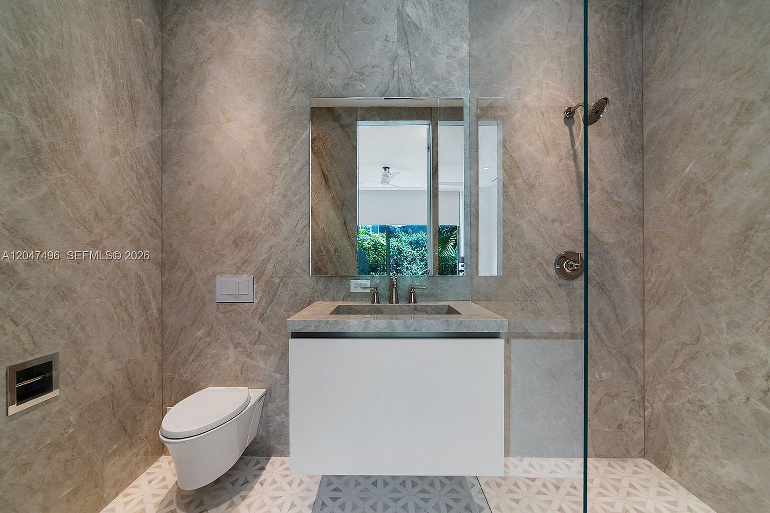 This modern bathroom features a wall-mounted toilet and a floating vanity with a stone countertop and integrated sink. The walls are clad in large-format, veined stone tiles, while the floor showcases a geometric patterned tile design. A glass shower partition separates the vanity area from the shower, creating a sleek and contemporary aesthetic.