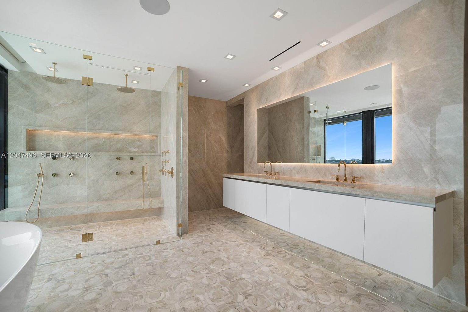 This luxurious primary bathroom features a spacious walk-in shower with glass walls and a large, wall-mounted vanity with dual gold-toned faucets. The room is finished with light-toned stone surfaces on the walls and a decorative, multi-textured stone floor, creating a sophisticated and cohesive aesthetic. A large, backlit mirror reflects the expansive window view, enhancing the bright and airy atmosphere of the space.