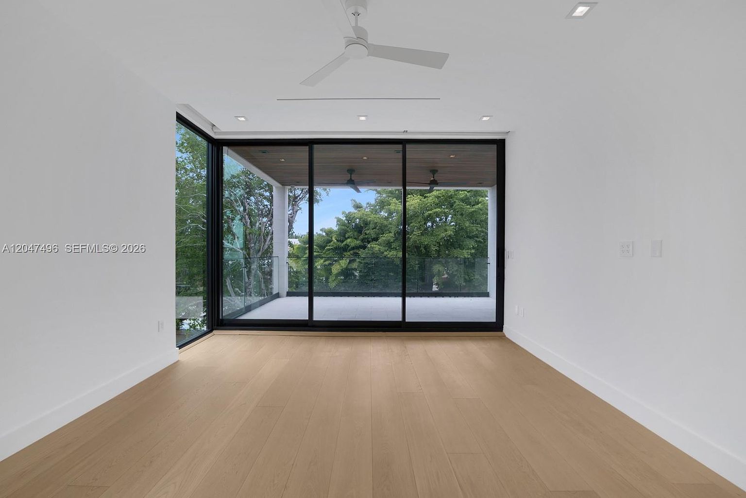 This minimalist living space features light wood flooring and crisp white walls, creating a bright and airy atmosphere. A large, corner-wrapping glass wall with sliding doors provides a seamless transition to a covered outdoor balcony, offering expansive views of the surrounding trees. The room is finished with a modern ceiling fan and recessed lighting, emphasizing a clean, contemporary aesthetic.