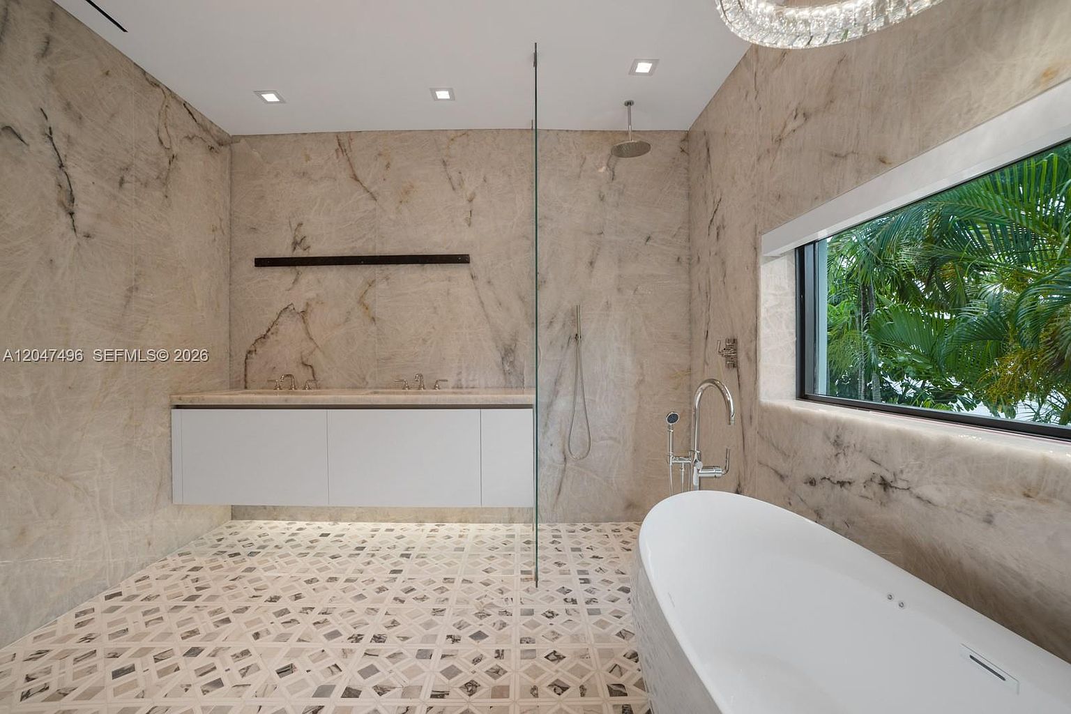 This luxurious primary bathroom features expansive stone wall cladding and a unique patterned tile floor that creates a sophisticated, high-end aesthetic. The space includes a sleek, wall-mounted vanity, a freestanding soaking tub positioned near a large window with lush greenery views, and a glass-enclosed shower area. The perspective captures the seamless integration of modern fixtures and natural textures, offering a serene and spa-like atmosphere.